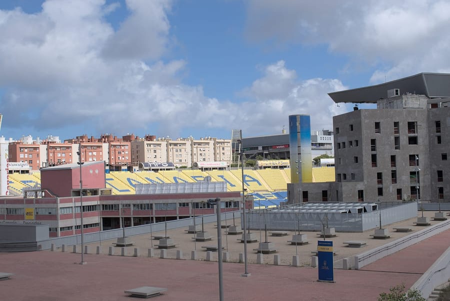 El Estadio de Gran Canaria visto desde la plaza exterior, con las gradas amarillas y azules formando la palabra CANARIA y los bloques de pisos del barrio al fondo.